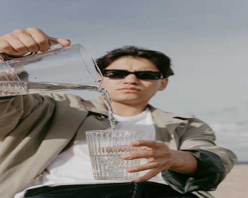 A person pouring cool clean water from a ceramic pitcher into a clear glass during a sunny afternoon break